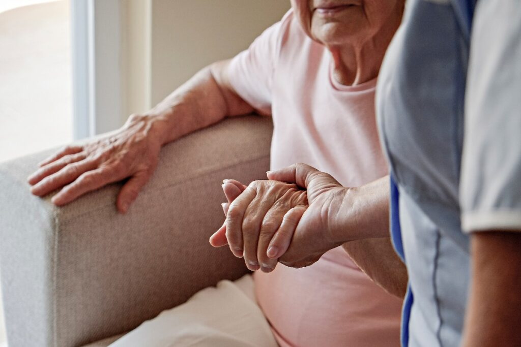 Caregiver supporta una donna anziana nell’alzarsi dalla poltrona, con primo piano sulle mani che si stringono.