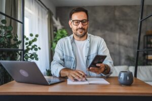 Uomo sorridente in ufficio mentre utilizza computer e cellulare